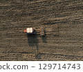 Red tractor plowing and cultivating soil in cultivated field, preparing for sowing, aerial view. 129714743