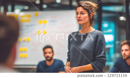 A confident businesswoman is leading a meeting, presenting ideas to her team in a modern office. She is standing and speaking passionately about a new project or strategy 129715159