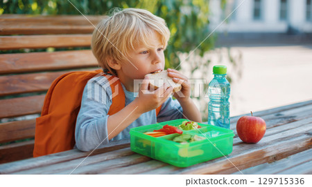 A young student with an orange backpack eats a sandwich while sitting at an outdoor picnic table. A bright green lunchbox with vegetables, a bottle of water, and an apple sit next to him 129715336