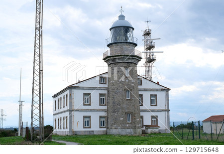 A beautiful historic lighthouse building in Asturias made of stone, standing majestically on a green hill with a grassy lawn. 129715648