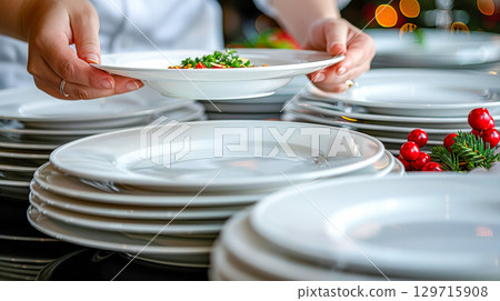 Hands serving a beautifully arranged plate of food from a stack of pristine white dinnerware 129715908