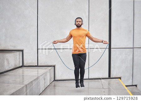Fitness, skipping rope and man jump in city for wellness exercise, cardio workout and training with headphones. Sports, energy and athlete jumping with gym equipment, listening to music in urban town 129715928