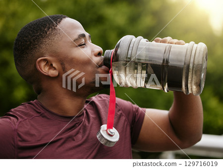 Fitness, nature and man drinking water after a workout in a park for hydration, thirst and rest. Sports, runner and healthy African male athlete enjoying a beverage during cardio exercise or training 129716019