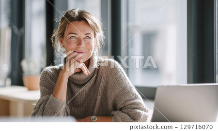 A middle-aged businesswoman is sitting at her desk in a modern office, smiling confidently at the camera. She is wearing a beige turtleneck sweater and a wristwatch. A laptop is next to her 129716070