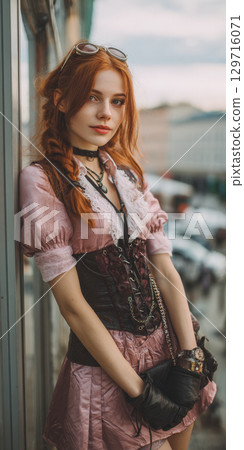 Woman with red hair is posing for photo. She is wearing a pink blouse, a brown corset, leather gloves, and steampunk sunglasses. The photo is taken on, city street, with cars, buildings in background. 129716071
