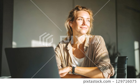 A smiling young woman sits at her home office desk with laptop, bathed in warm sunlight. This image captures essence of successful freelancer or remote worker enjoying her career and work-life balance 129716075