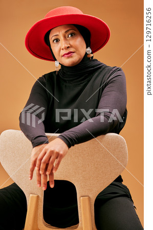Fashion, beauty and muslim woman on chair with stylish hat, cosmetics and trendy makeup on studio background. Mature model portrait, islamic hijab and culture, lifestyle or luxury clothes in Malaysia 129716256