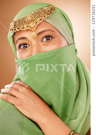 Fashion, tradition and face of a muslim woman in studio with traditional jewelry and burka. Beauty, islam and portrait of mature lady with islamic hijab scarf or arabic jewellery by brown background. 129716281