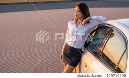 A woman leaning against her car in a parking lot at dusk, gazing into the distance 129716296