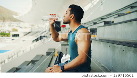 Stadium, man and drink water with earphones for fitness, hydration and running break. Outdoor, athlete person and relax with liquid beverage for training podcast, exercise and music for workout rest Stadium, man and drink water with earphones for fitness, hydration and running break. Outdoor, athlete person and relax with liquid beverage for training podcast, exercise and music for workout rest 129716501