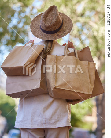 Sun, trees and woman with bags from shopping from back walking in park on holiday. Summer, nature and travel, girl with shopping bag from luxury designer boutique buying gifts on vacation garden path 129717434