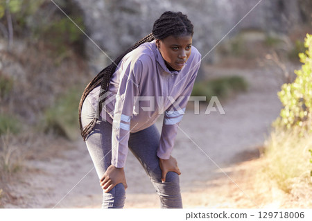 Fitness, mountain and black woman tired from running, marathon training and exercise in nature. Wellness, endurance and female athlete taking a break, rest and breathe to relax after sports workout 129718006