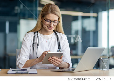 Smiling female doctor using tablet in office. Wearing white coat, stethoscope, glasses, working on laptop. Represents healthcare, technology, professionalism, medical practice. 129718007