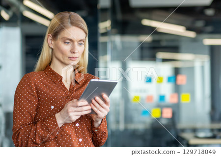 A focused businesswoman in an office setting, holding a tablet, appears engrossed in her work. The modern office has glass walls. A focused businesswoman in an office setting, holding a tablet, appears engrossed in her work. The modern office has glass walls. 129718049