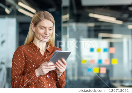 A businesswoman with blonde hair smiles as she holds a tablet in an office setting, possibly working or communicating. 129718051