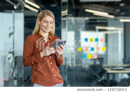 A blonde businesswoman uses a tablet inside a modern office, with glass walls and colorful sticky notes adding a vibrant touch. 129718053