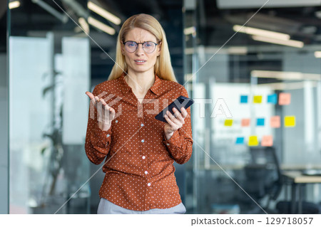 A frustrated woman in glasses holds a phone, gesturing with confusion in an office setting with glass walls and colorful sticky notes. A frustrated woman in glasses holds a phone, gesturing with confusion in an office setting with glass walls and colorful sticky notes. 129718057