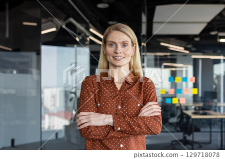 Smiling businesswoman standing with arms crossed in modern office. Professional demeanor exudes confidence and leadership. Ideal representation of success and empowerment in workplace. 129718078