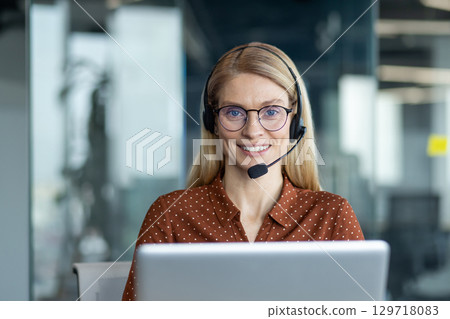 A friendly blonde woman with glasses and a headset smiles at the camera while working on her laptop in a modern office. A friendly blonde woman with glasses and a headset smiles at the camera while working on her laptop in a modern office. 129718083
