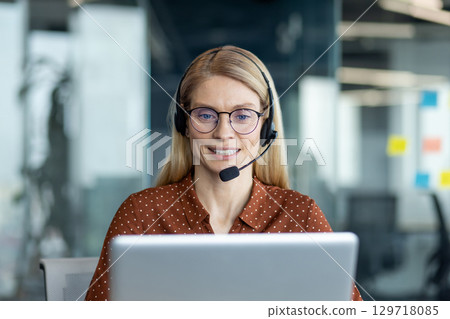 A friendly customer service representative smiles while wearing a headset and working on a laptop at a modern office. 129718085