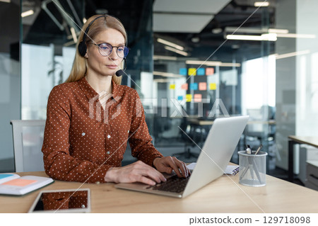 A businesswoman wearing a headset works at her laptop in a modern office environment. She is focused and engaged in her work with a serious expression. A businesswoman wearing a headset works at her laptop in a modern office environment. She is focused and engaged in her work with a serious expression. 129718098