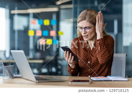 Businesswoman expressing frustration while looking at phone during work. Scene captures emotional response, phone technology, and office setting. Includes laptop and notes on desk. 129718125