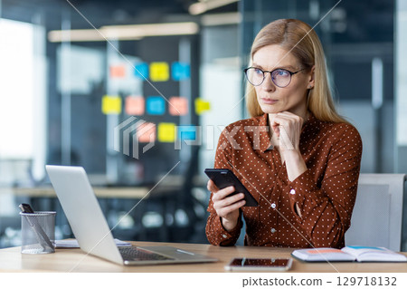 Professional woman in business attire using smartphone and laptop at office desk. Concentrated on planning and decision-making, exploring technology for efficient work. Professional woman in business attire using smartphone and laptop at office desk. Concentrated on planning and decision-making, exploring technology for efficient work. 129718132