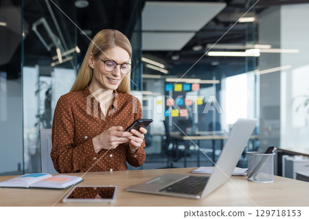 Joyful businesswoman using phone while sitting inside office, woman typing message using app on smartphone, working with laptop inside office. 129718153