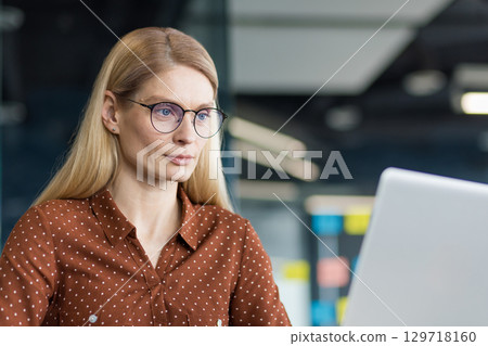 Mature woman close-up at workplace working with laptop, businesswoman looking at computer screen while sitting at desk, confident and concentrated. Mature woman close-up at workplace working with laptop, businesswoman looking at computer screen while sitting at desk, confident and concentrated. 129718160