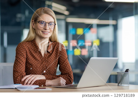 Portrait of a successful businesswoman at her workplace inside the office. Office worker smiling and looking at the camera, sitting at a desk with a laptop. Portrait of a successful businesswoman at her workplace inside the office. Office worker smiling and looking at the camera, sitting at a desk with a laptop. 129718189