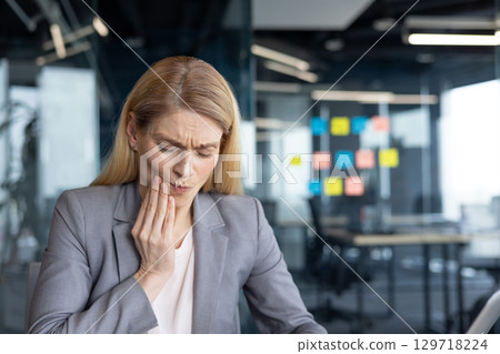Mature woman businesswoman holding her jaw in pain at work in an office. Depicting discomfort possibly from a dental issue, this conveys feelings of distress and the need for healthcare attention. 129718224