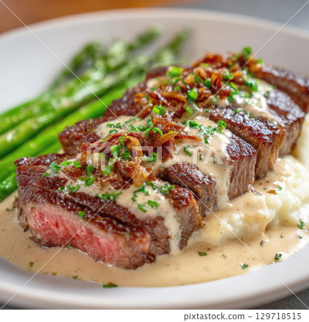A gourmet steak dinner is beautifully plated with sliced steak on mashed potatoes, topped with sauce and fried onions. Dish includes asparagus, highlighting high-end dining, professional culinary art 129718515