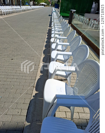 White plastic chairs are neatly arranged along the cobblestone streets in Spain, preparing for a vibrant parade. People will soon gather to enjoy the festivities and celebrations. White plastic chairs are neatly arranged along the cobblestone streets in Spain, preparing for a vibrant parade. People will soon gather to enjoy the festivities and celebrations. 129718925