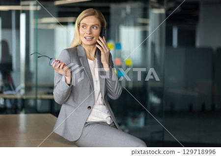 Smiling businesswoman in gray suit engaged in phone conversation in modern office environment. Holding eyeglasses, showcasing confidence and professionalism in business setting. Smiling businesswoman in gray suit engaged in phone conversation in modern office environment. Holding eyeglasses, showcasing confidence and professionalism in business setting. 129718996