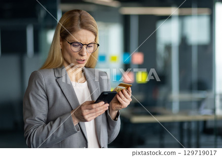 Businesswoman with glasses in office holding credit card and phone, looking concerned. Modern technology use, possible online payment issue or identity theft concept. Businesswoman with glasses in office holding credit card and phone, looking concerned. Modern technology use, possible online payment issue or identity theft concept. 129719017