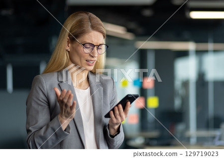 Businesswoman in a gray suit expressing frustration while using smartphone in office. Glasses, suit, and expressive gestures illustrate work stress or communication issues. Businesswoman in a gray suit expressing frustration while using smartphone in office. Glasses, suit, and expressive gestures illustrate work stress or communication issues. 129719023