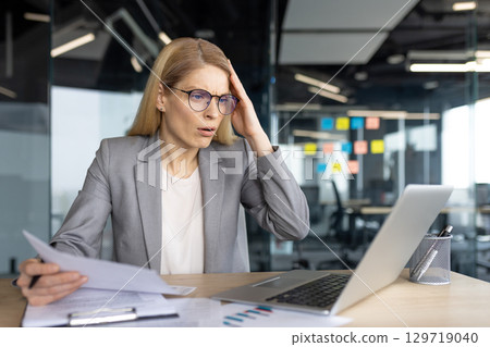 A businesswoman wearing a suit looks concerned while reviewing documents at her desk in an office. 129719040