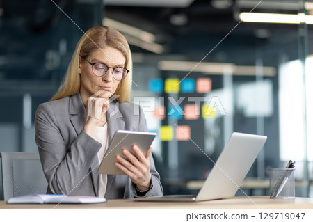 Professional adult woman focused on reading a tablet in an office with modern decor. 129719047