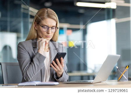 A professional woman using a smartphone while seated at her desk in a modern office. The scene conveys focus, determination, and efficiency in a business or corporate workspace. 129719061