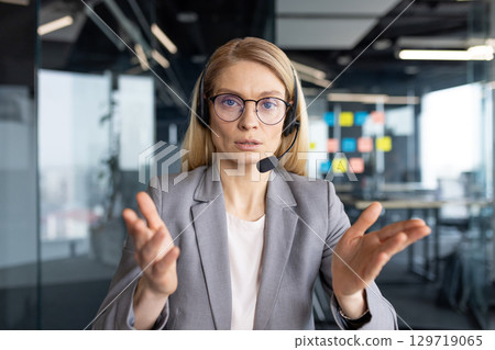 Serious businesswoman with headset communicating during online video call, view from webcam. Concept of remote work, virtual meeting, customer support, and digital communication. 129719065