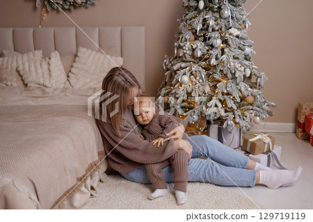 Young mother holding baby near decorated Christmas tree in cozy bedroom, front view Young mother holding baby near decorated Christmas tree in cozy bedroom, front view 129719119