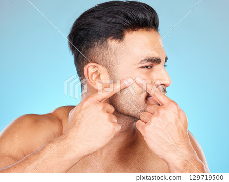 Man, hands and pop acne of face for skincare, beauty wellness or skin dermatology. Indian man, upset and press facial pimples or blackhead in blue background studio for morning cosmetics care Man, hands and pop acne of face for skincare, beauty wellness or skin dermatology. Indian man, upset and press facial pimples or blackhead in blue background studio for morning cosmetics care 129719500