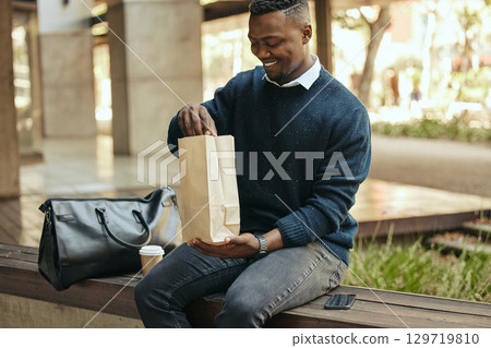 Office employee with a coffee, a paper sandwich bag and a smile, on a bench for a break from his busy work schedule. Happy, hungry businessman, time to eat, with his food and drink outside for lunch. Office employee with a coffee, a paper sandwich bag and a smile, on a bench for a break from his busy work schedule. Happy, hungry businessman, time to eat, with his food and drink outside for lunch. 129719810