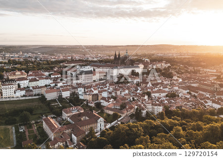 Golden sunlight bathes Prague Castle and the surrounding city at dawn. The vibrant rooftops glow as the first light breaks, showcasing a breathtaking panorama of historical architecture. 129720154
