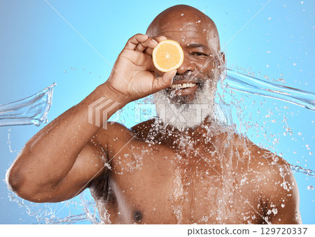 Man face, fruit or water splash on blue background in studio for vitamin c treatment, healthcare wellness or cleaning hygiene. Portrait, smile happy or mature skincare for black model with wet lemon 129720337