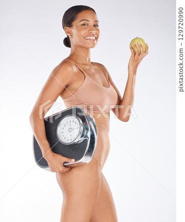 Portrait, black woman and body with apple, scale and diet for weight loss, health and wellness on grey studio background. Face, African American female and happy lady with fruit, fitness or nutrition 129720990