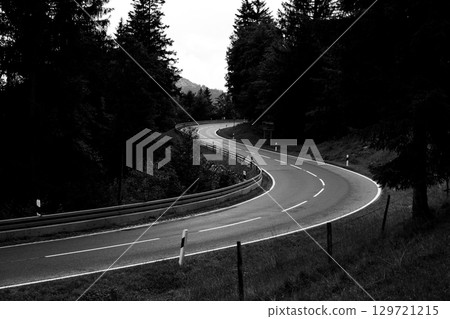 A winding road gently curves through a dense forest, surrounded by trees and greenery. The ambiance displays a tranquil yet mysterious atmosphere, suggesting calm driving conditions. A winding road gently curves through a dense forest, surrounded by trees and greenery. The ambiance displays a tranquil yet mysterious atmosphere, suggesting calm driving conditions. 129721215