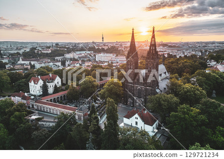 Morning light illuminates Vysehrad Basilica of St. Peter and St. Paul, showcasing its stunning architecture against a backdrop of Prague's skyline and lush greenery. 129721234