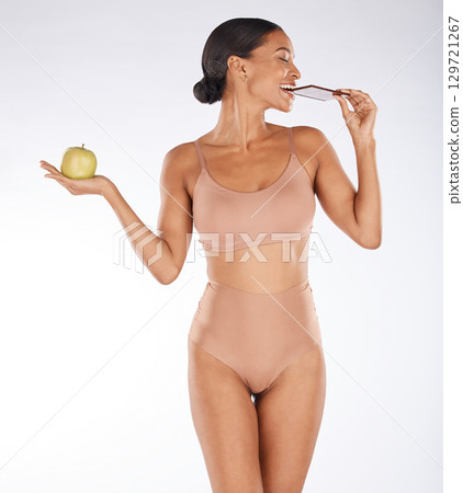Apple, body and woman eating chocolate in a studio for a sweet snack, candy or craving. Health, wellness and female model enjoying cocoa treat while holding fruit for balance diet by gray background. Apple, body and woman eating chocolate in a studio for a sweet snack, candy or craving. Health, wellness and female model enjoying cocoa treat while holding fruit for balance diet by gray background. 129721267