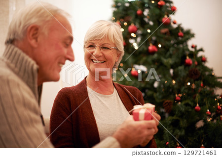 Senior couple, giving gift and together for Christmas holiday celebration at home with a smile, happiness and kindness. Surprise, celebrate and tree with man and woman holding a present and gift box 129721461